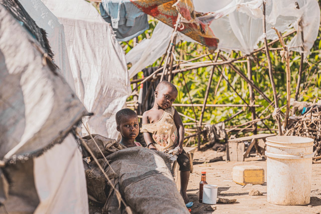 Two children sitting by makeshift shelters in a rural village, surrounded by nature.